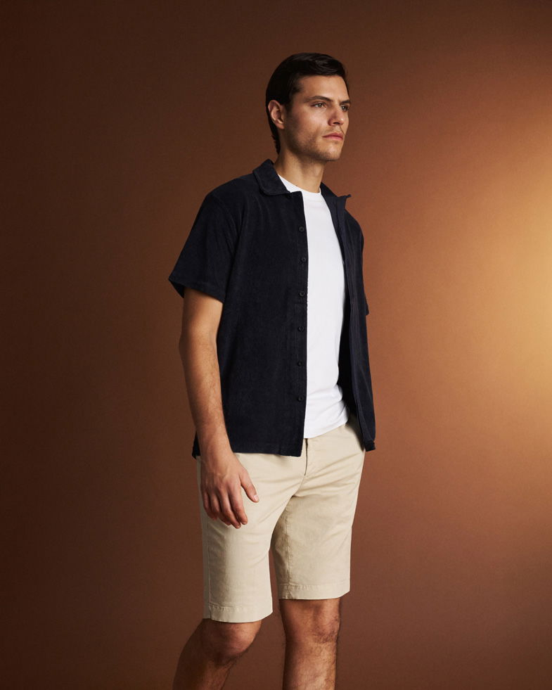 Mann steht vor braunem Hintergrund, trägt beige Shorts, weißes T-Shirt und offenes, dunkelblaues Kurzarmhemd. Mann steht vor braunem Hintergrund, trägt beige Shorts, weißes T-Shirt und offenes, dunkelblaues Kurzarmhemd.