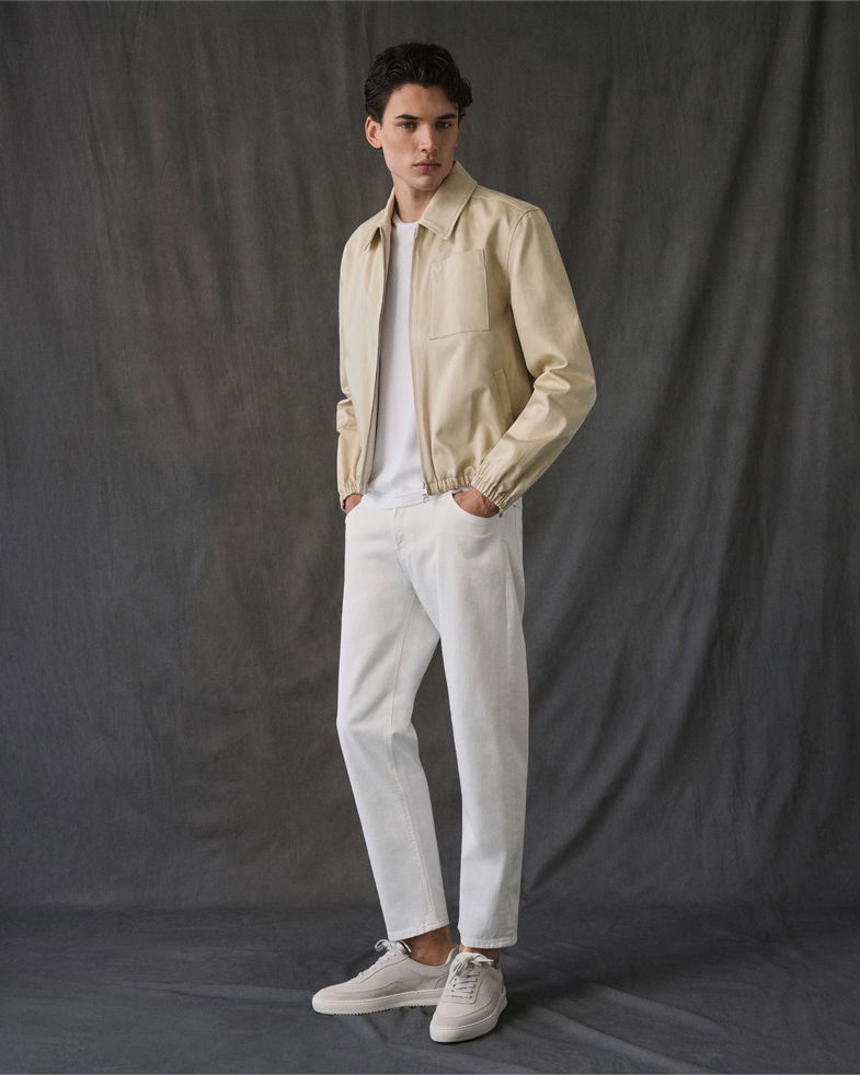 Mann trägt beige Jacke, weißes T-Shirt, weiße Slim-Fit-Jeans und beige Sneaker vor grauem Hintergrund.