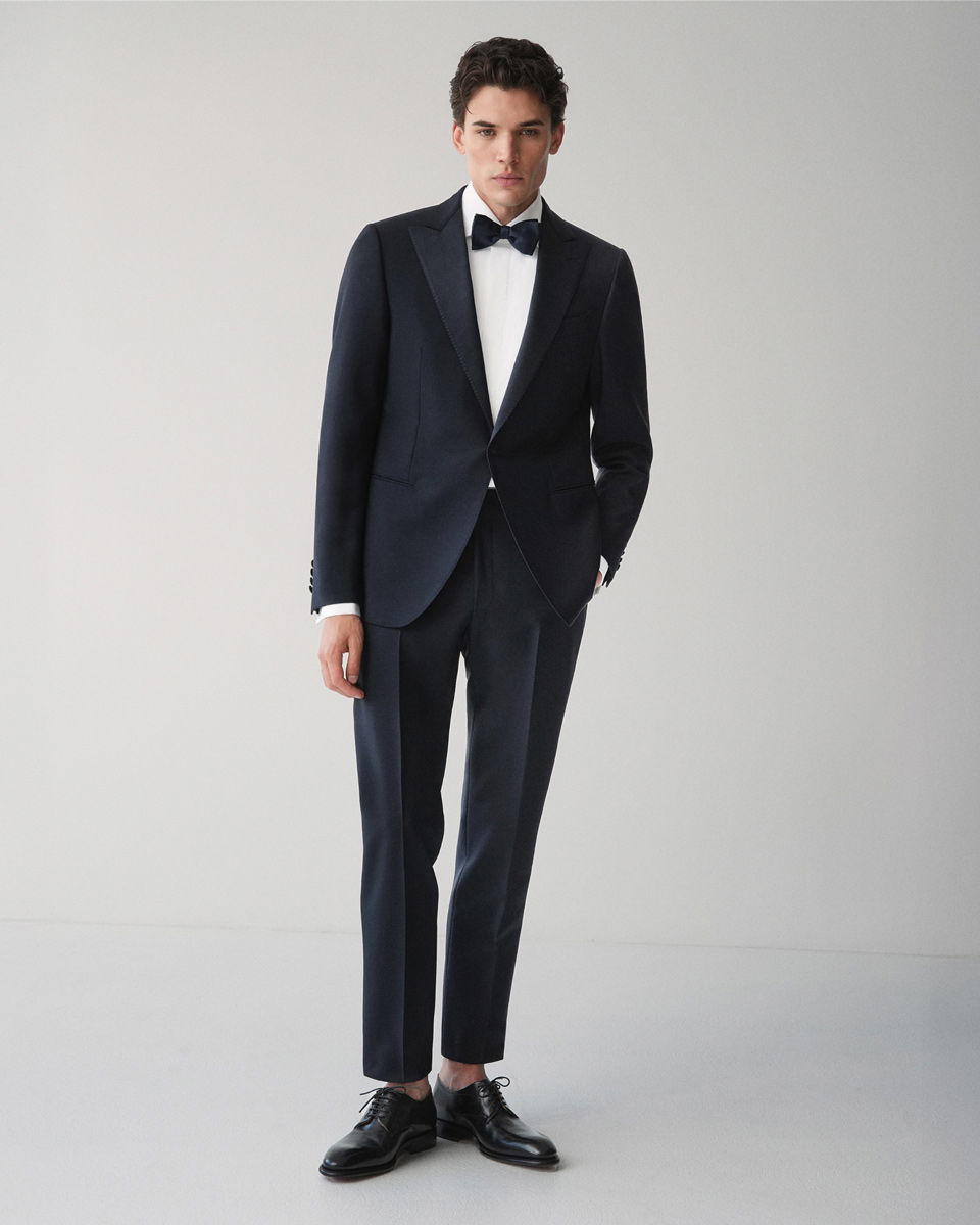 Mann in schwarzem Smoking mit spitzem Revers, weißem Hemd, schwarzer Fliege und schwarzen Lederschuhen vor hellem Hintergrund.