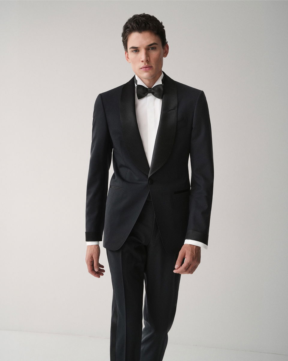 Junger Mann mit schwarzem Smoking, weißem Hemd und schwarzer Fliege vor hellem Hintergrund stehend.