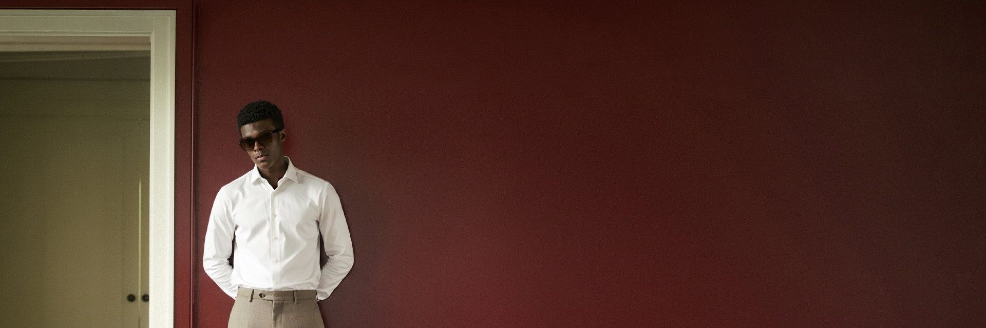 Mann mit dunkler Haut trägt weiße Hemd, beige Hose und schwarze Sonnenbrille, lehnt vor bordeauxroter Wand neben Türrahmen. Mann mit dunkler Haut trägt weiße Hemd, beige Hose und schwarze Sonnenbrille, lehnt vor bordeauxroter Wand neben Türrahmen.