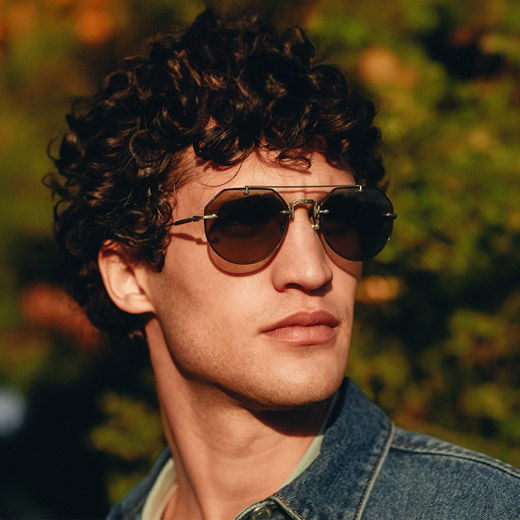 Junger Mann mit lockigem Haar trägt braun-goldene Aviator-Sonnenbrille mit Titanrahmen und Jeansjacke vor grünem Hintergrund.