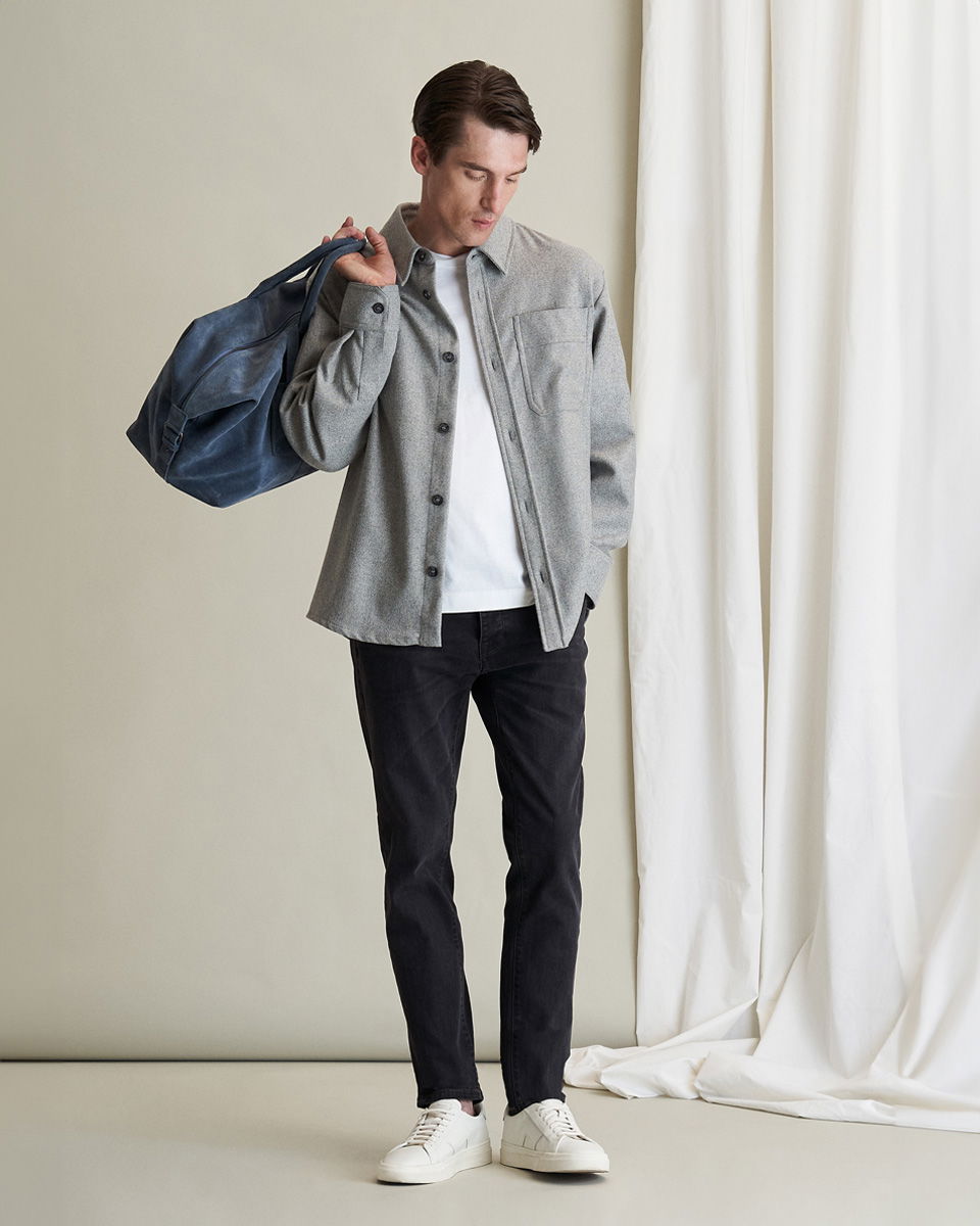 Mann trägt graues Overhemd, weißes T-Shirt, schwarze Hose, weiße Sneaker und trägt blauen Weekender über der Schulter. Mann trägt graues Overhemd, weißes T-Shirt, schwarze Hose, weiße Sneaker und trägt blauen Weekender über der Schulter.