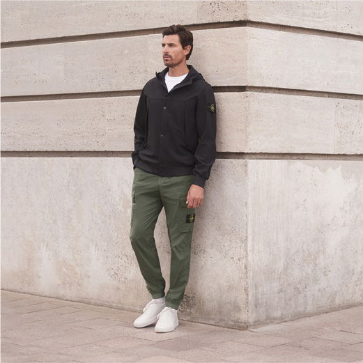 Mann lehnt an Hausecke, trägt weiße Sneaker, olivgrüne Cargohose, weißes T-Shirt und schwarze Jacke mit Kapuze. Mann lehnt an Hausecke, trägt weiße Sneaker, olivgrüne Cargohose, weißes T-Shirt und schwarze Jacke mit Kapuze.