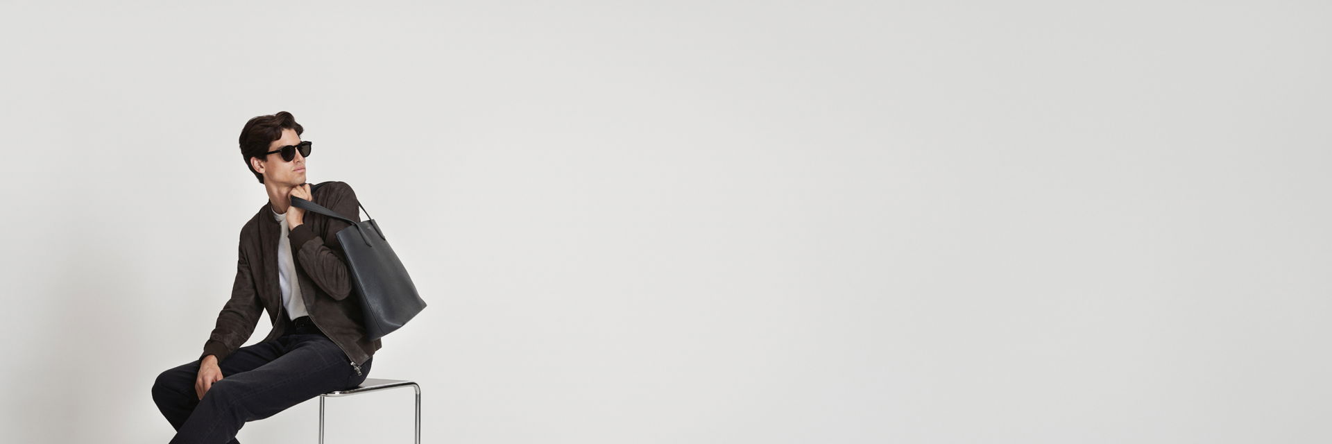 Mann mit braunem Veloursleder-Blouson, weißem Shirt, schwarzer Jeans, schwarzer Tote-Bag und Sonnenbrille sitzt auf Hocker. Mann mit braunem Veloursleder-Blouson, weißem Shirt, schwarzer Jeans, schwarzer Tote-Bag und Sonnenbrille sitzt auf Hocker.
