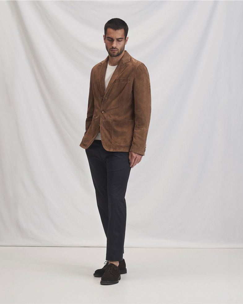 Mann trägt braunen Veloursleder-Blazer, beige Langarmshirt, schwarze Hose und braune Loafer vor weißem Hintergrund.