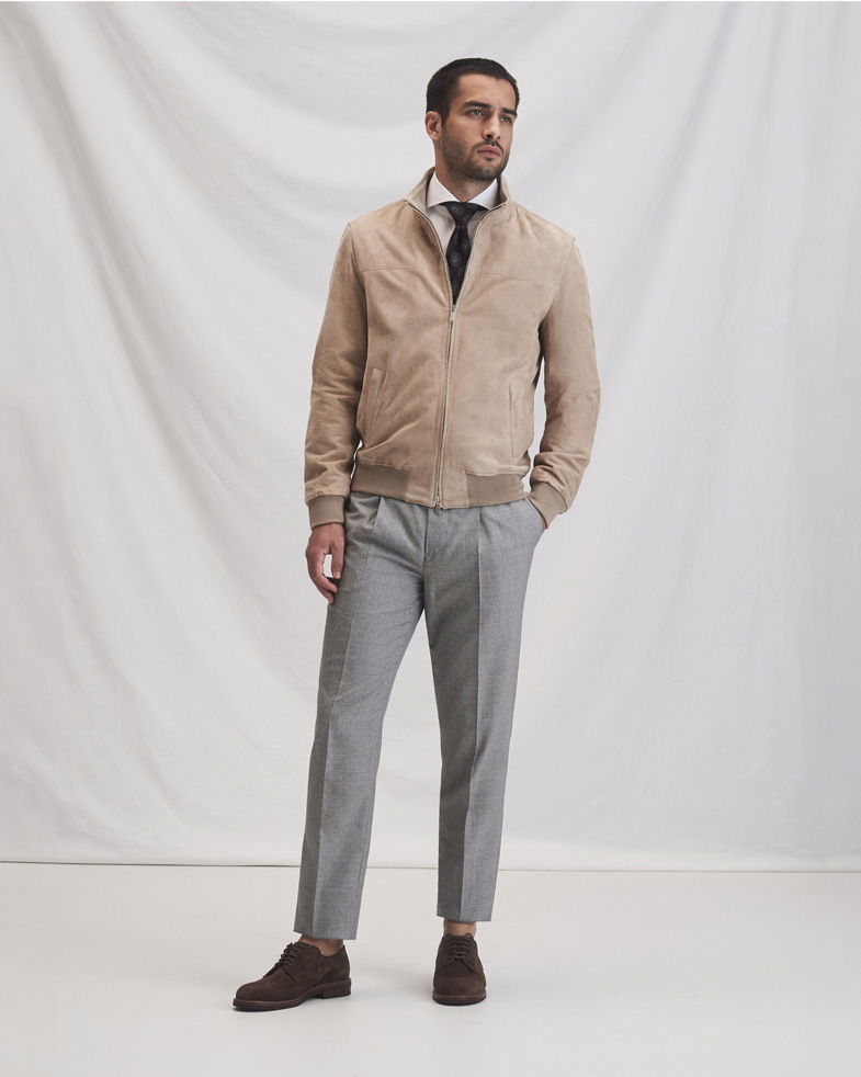 Mann trägt beige Veloursleder-Blouson, weißes Hemd mit schwarzer Krawatte, graue Hose und braune Schnürschuhe vor weißem Hintergrund.