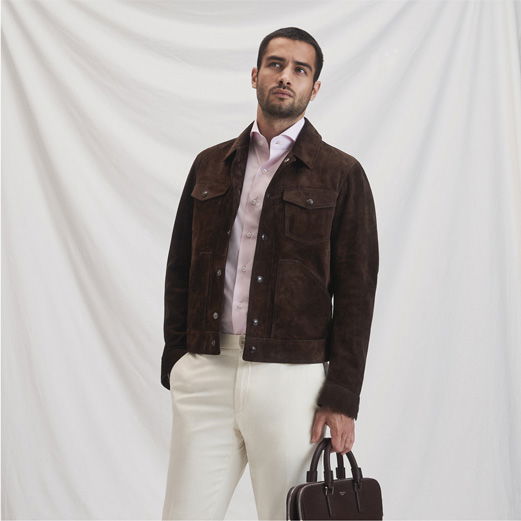 Mann trägt dunkelbraune Velourslederjacke, rosa Hemd, weiße Hose und hält schwarze Aktentasche vor weißem Hintergrund.