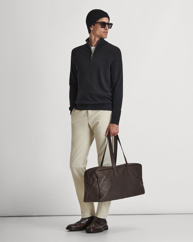 Mann mit schwarzem Troyer, beiger Hose, schwarzer Mütze, Sonnenbrille, braunen Schnürschuhen hält dunkelbraunen Weekender. Mann mit schwarzem Troyer, beiger Hose, schwarzer Mütze, Sonnenbrille, braunen Schnürschuhen hält dunkelbraunen Weekender.