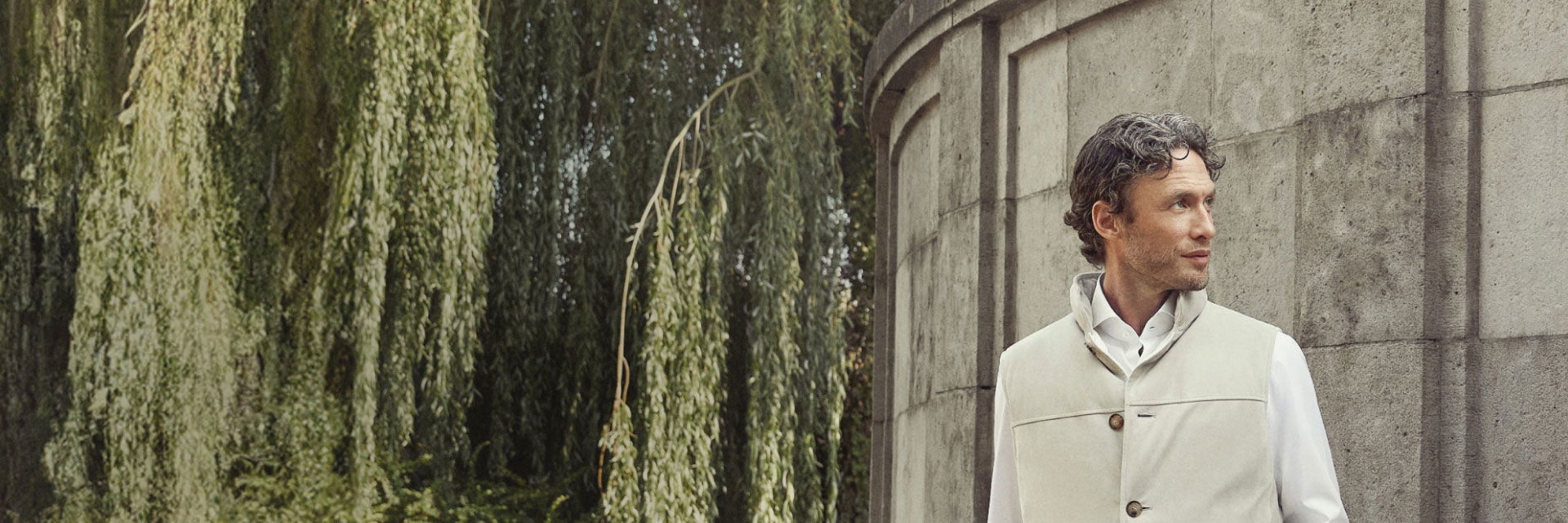 Mann mit beige Weste über weißem Hemd lehnt an grauer Mauer, im Hintergrund hängende grüne Weidenzweige, Blick nach rechts. Mann mit beige Weste über weißem Hemd lehnt an grauer Mauer, im Hintergrund hängende grüne Weidenzweige, Blick nach rechts.