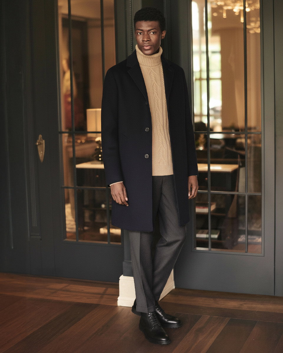 Mann in beige Zopfpullover, dunkelgrauer Hose, navy Mantel und schwarzen Schnürboots steht vor Glastür in elegantem Raum. Mann in beige Zopfpullover, dunkelgrauer Hose, navy Mantel und schwarzen Schnürboots steht vor Glastür in elegantem Raum.