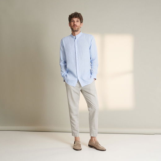 Mann mit kurzem braunem Haar trägt hellblaues Hemd, beige Hose und beige Loafers, steht vor hellem Hintergrund.