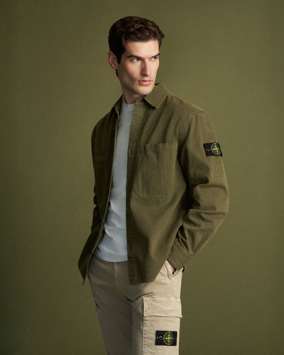 Mann trägt olivfarbenes Overshirt über weißem T-Shirt und beigefarbene Cargohose, Blick nach links, Hände in Taschen.