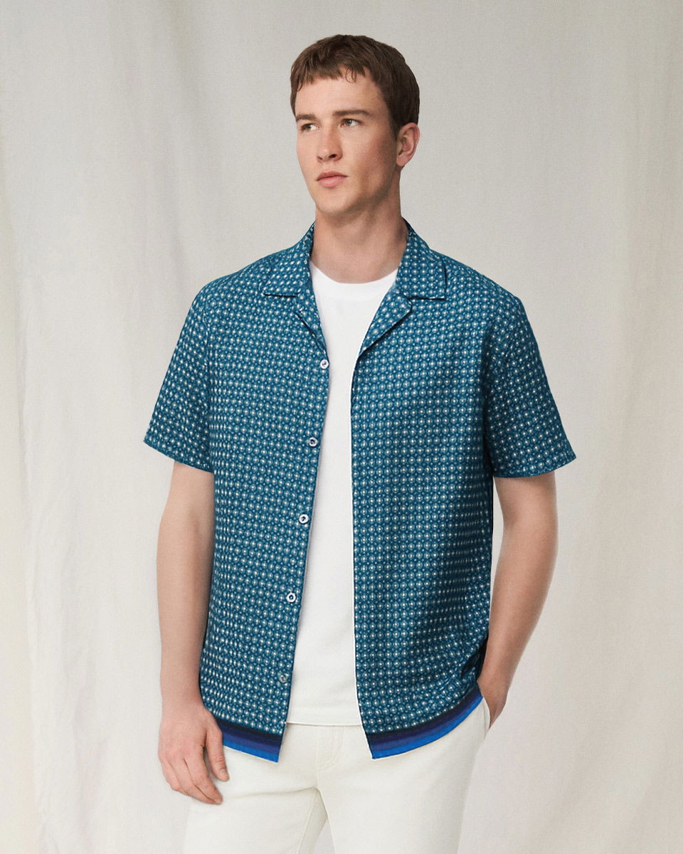 Mann trägt blau gemustertes Kurzarmhemd über weißem T-Shirt und weiße Hose, schaut nach links, steht vor hellem Hintergrund. Mann trägt blau gemustertes Kurzarmhemd über weißem T-Shirt und weiße Hose, schaut nach links, steht vor hellem Hintergrund.