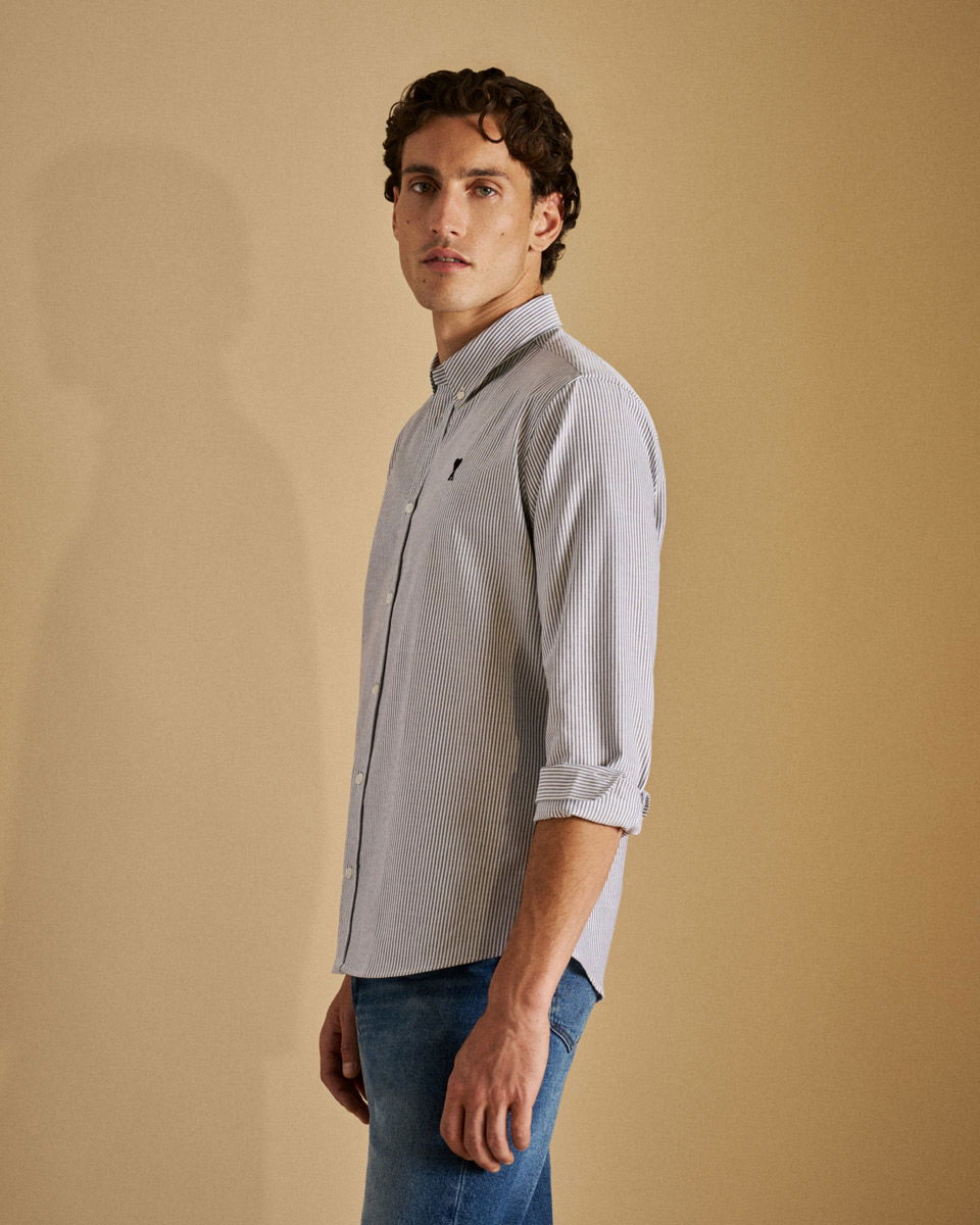 Mann mit lockigem Haar trägt blau-weiß gestreiftes Hemd mit italienischer Krempelung und blaue Jeans vor beigem Hintergrund.