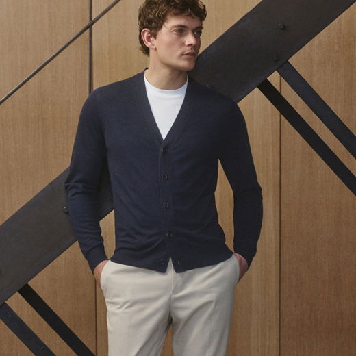 Mann mit dunkelblauem Cardigan, weißem T-Shirt und beiger Chino lehnt vor einer Holz- und Metalltreppe, Hände in Taschen.