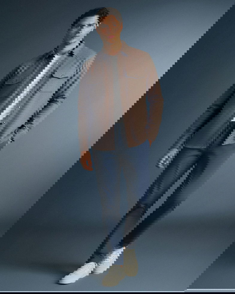 Mann trägt blaue Chinohose, weißes T-Shirt, braunes Overshirt und weiße Sneaker vor grauem Hintergrund.