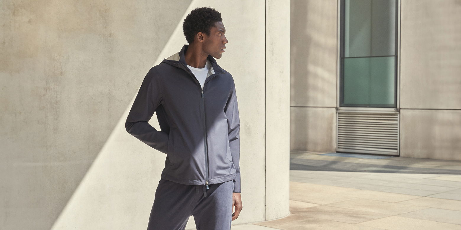 Mann in dunkelblauer Windbreaker-Jacke, weißem T-Shirt und blauer Hose steht vor hellem Betongebäude und schaut nach links. Mann in dunkelblauer Windbreaker-Jacke, weißem T-Shirt und blauer Hose steht vor hellem Betongebäude und schaut nach links.