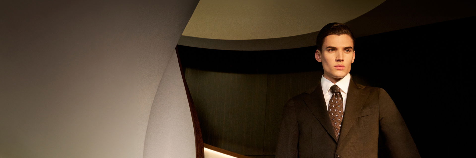 Junger Mann in braunem Sakko, weißem Hemd und braun gemusterter Krawatte vor modernem, dunklem Hintergrund stehend. Junger Mann in braunem Sakko, weißem Hemd und braun gemusterter Krawatte vor modernem, dunklem Hintergrund stehend.