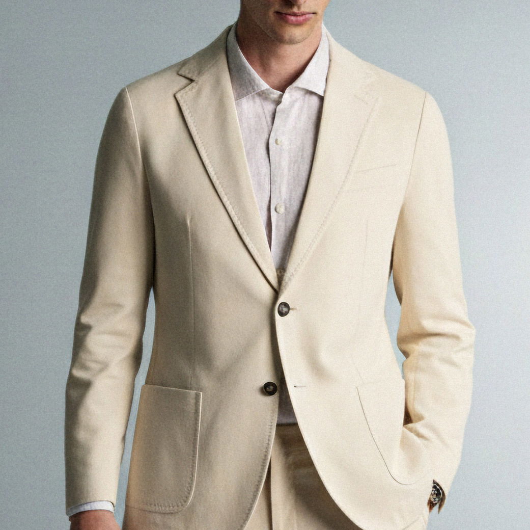 Herrenanzug in Creme mit aufgesetzten Taschen, kombiniert mit hellgrauem Hemd, Manschette mit Uhr sichtbar, Oberkörper frontal.