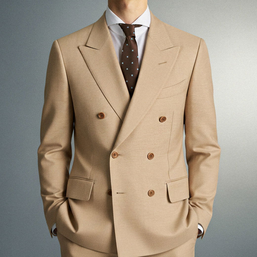 Herr trägt beige doppelreihigen Anzug mit brauner gemusterter Krawatte und weißem Hemd, Hände in den Hosentaschen.