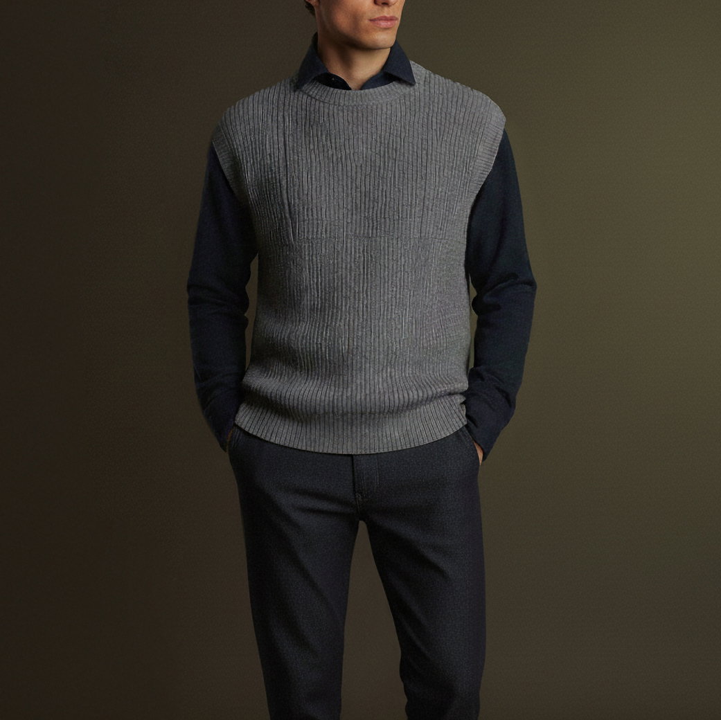Mann trägt dunkelblaues Hemd mit grauem, strukturiertem Pullunder und dunkelgraue Hose, Hände in Taschen, vor neutralem Hintergrund.