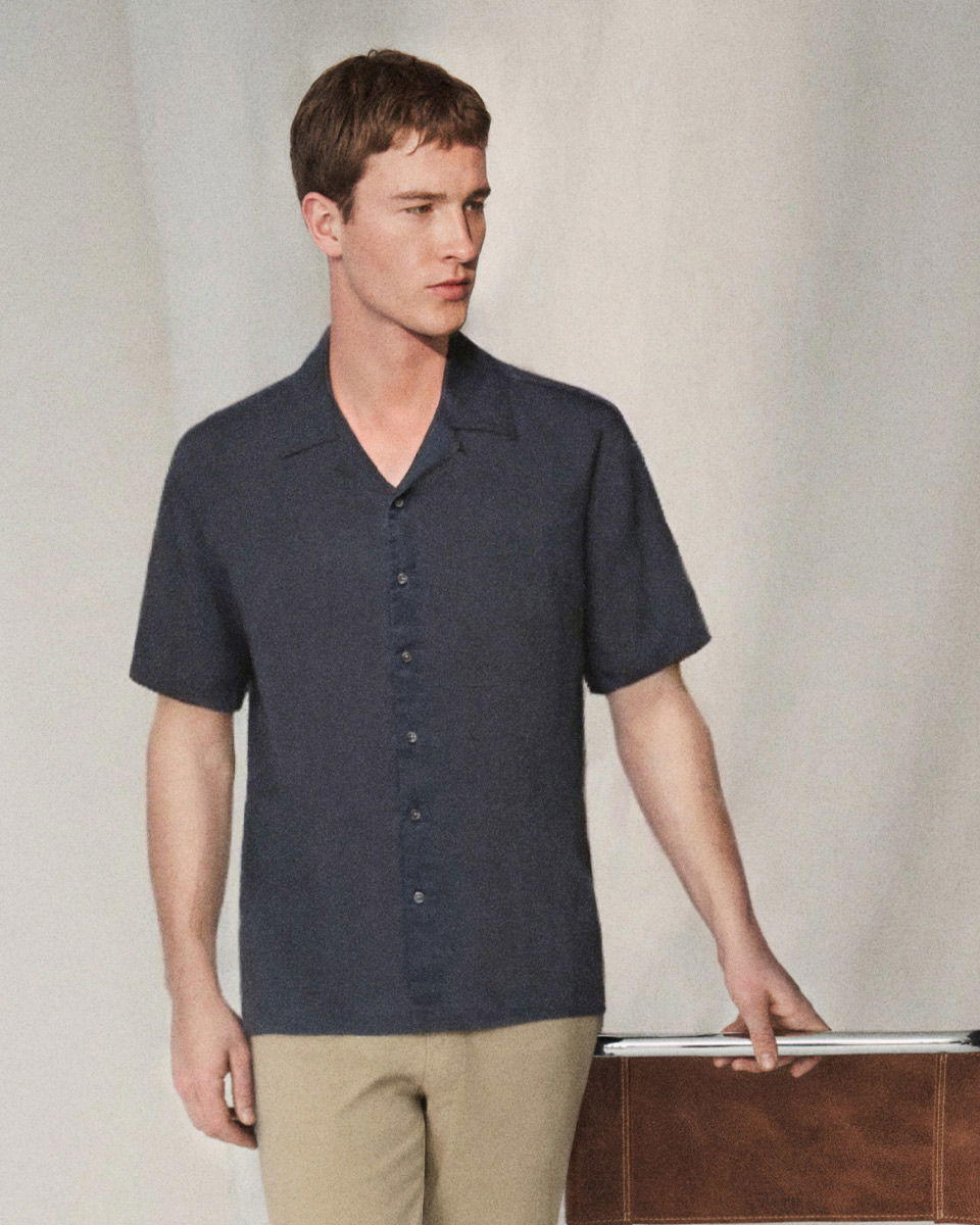 Mann mit kurz geschnittenem braunem Haar trägt dunkelblaues Kurzarmhemd und beigefarbene Hose, lehnt an brauner Ledertasche. Mann mit kurz geschnittenem braunem Haar trägt dunkelblaues Kurzarmhemd und beigefarbene Hose, lehnt an brauner Ledertasche.