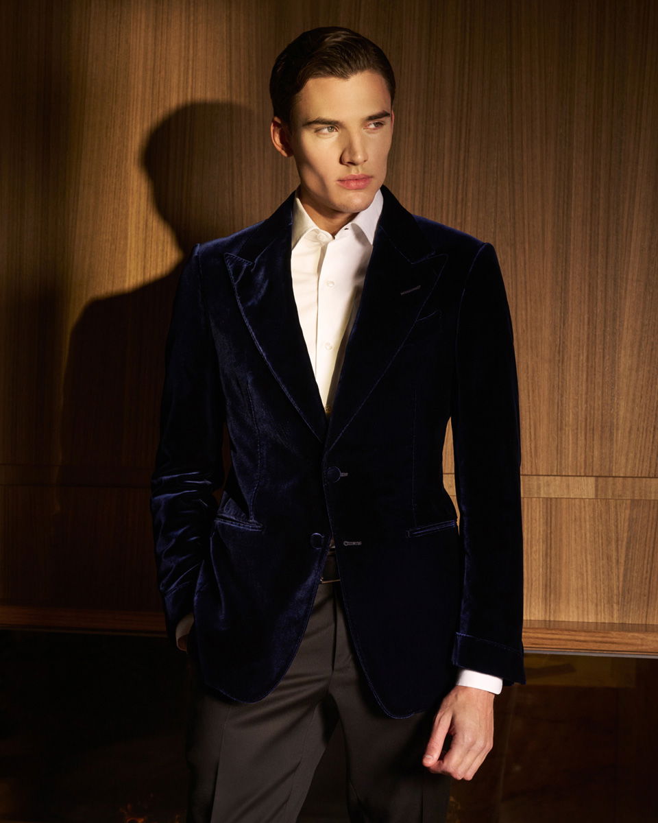 Mann mit dunkelblauem Samtsakko, weißem Hemd und schwarzer Hose steht vor einer Holzpaneelwand, schaut nach rechts. Mann mit dunkelblauem Samtsakko, weißem Hemd und schwarzer Hose steht vor einer Holzpaneelwand, schaut nach rechts.