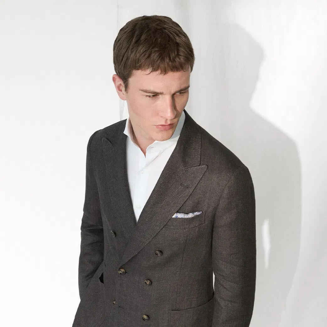 Mann mit dunkelgrauem, doppelreihigem Sakko, weißem Hemd und gemustertem Einstecktuch steht vor weißer Wand und schaut nach links.