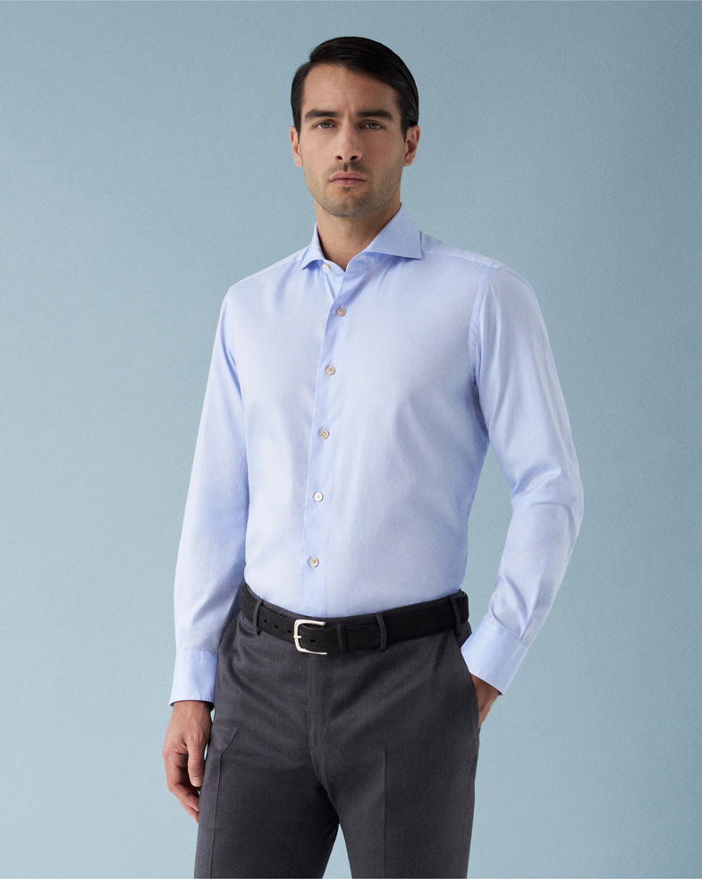 Mann mit dunkelgrauer Hose, hellblauem Hemd und schwarzem Gürtel vor einfarbig hellblauem Hintergrund.