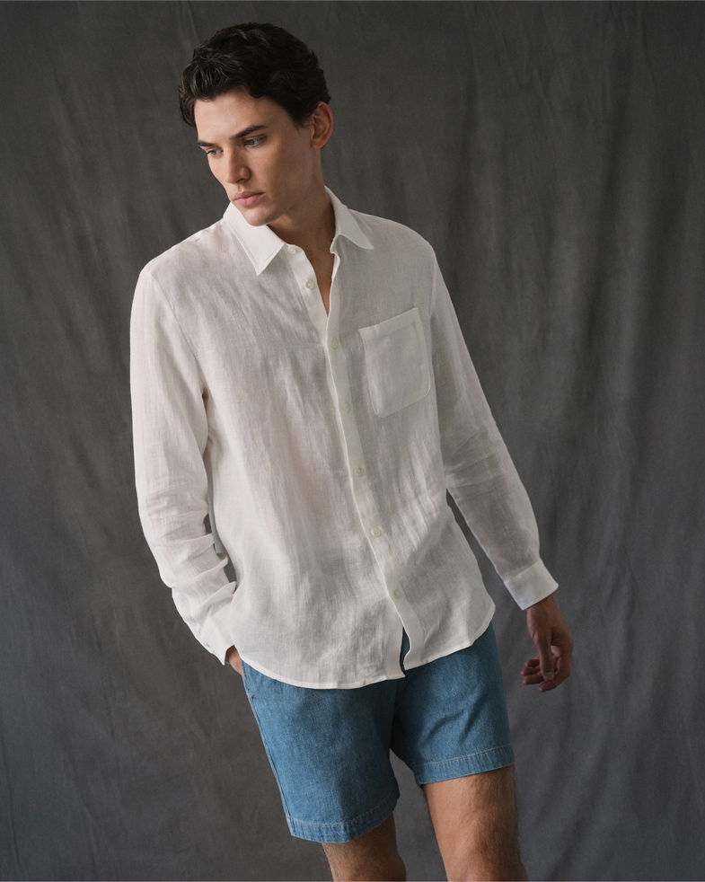 Junger Mann trägt weißes Leinenhemd mit Brusttasche und hellblaue Jeansshorts vor grauem Stoffhintergrund.