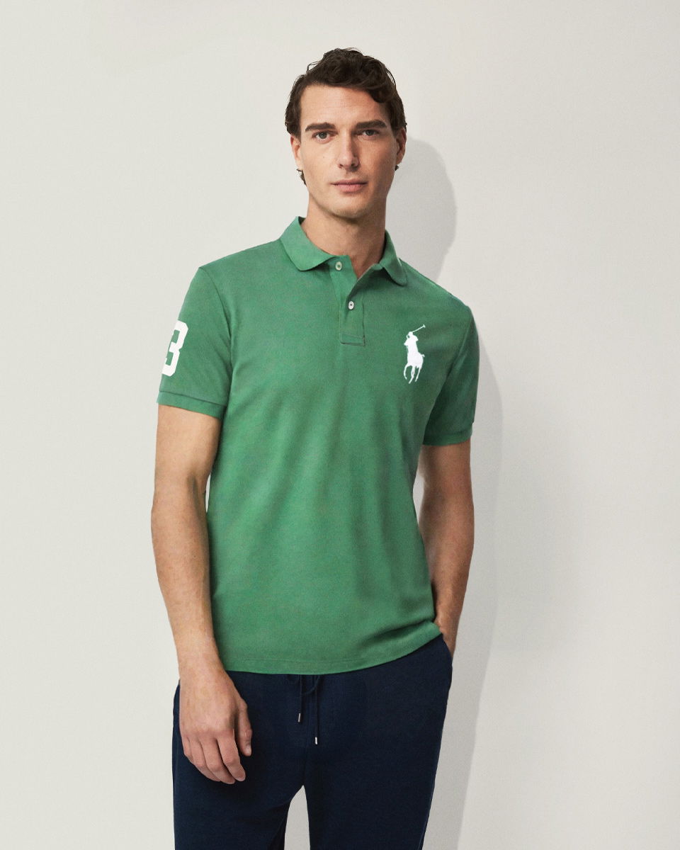 Mann mit dunkelblauer Jogpants und grünem Poloshirt mit großem weißem Designerlogo und Zahl 3 am Ärmel vor weißem Hintergrund.