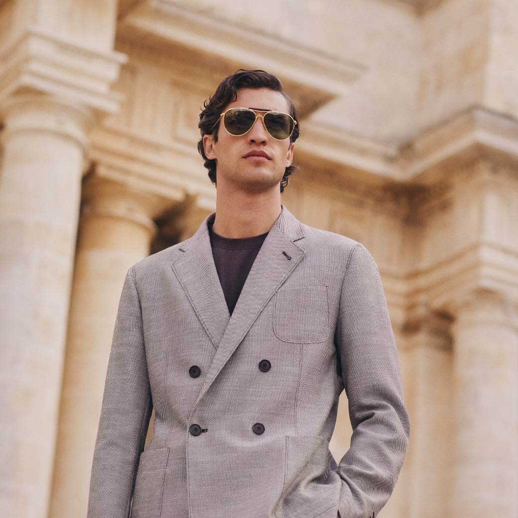 Mann mit grauem, gemustertem Doppelreiher-Sakko, schwarzem Rundhalsshirt und goldener Sonnenbrille vor Säulengebäude. Mann mit grauem, gemustertem Doppelreiher-Sakko, schwarzem Rundhalsshirt und goldener Sonnenbrille vor Säulengebäude.
