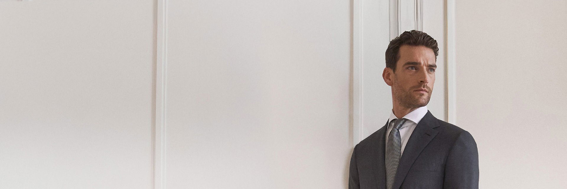 Herr mit dunklem Anzug, weißem Hemd und gemusterter Krawatte steht vor heller Wand und blickt nach rechts. Herr mit dunklem Anzug, weißem Hemd und gemusterter Krawatte steht vor heller Wand und blickt nach rechts.