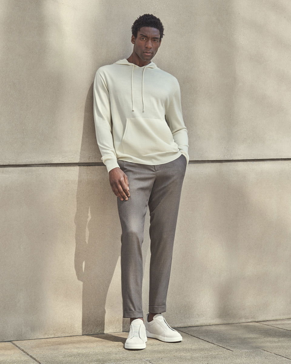 Mann lehnt an Betonwand, trägt cremefarbenen Hoodie, graue Hose und weiße Sneaker, steht auf Pflastersteinboden. Mann lehnt an Betonwand, trägt cremefarbenen Hoodie, graue Hose und weiße Sneaker, steht auf Pflastersteinboden.