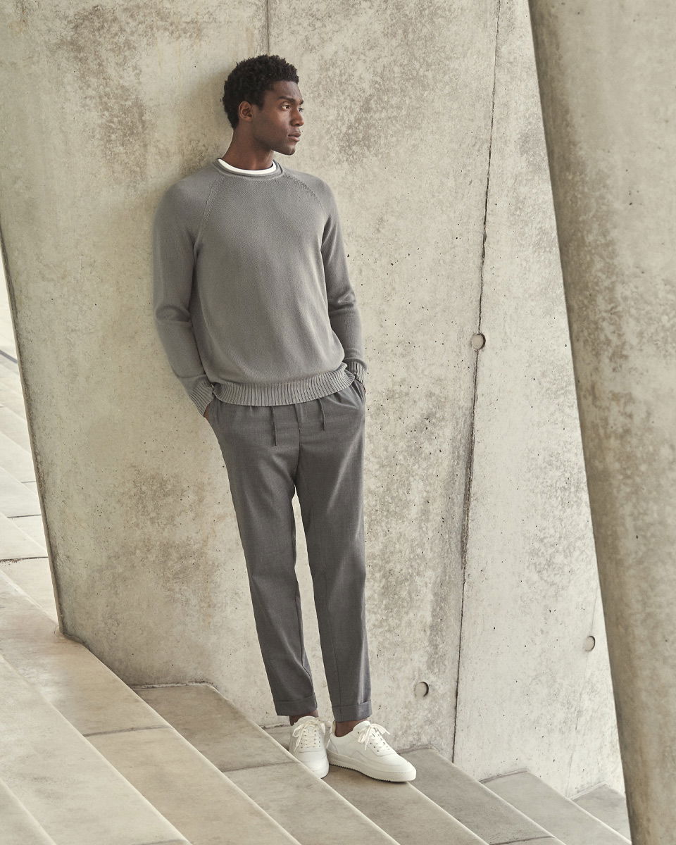 Mann mit grauem Rundhalspullover, grauer Hose und weißen Sneakern lehnt entspannt an Betonwand auf Treppe. Mann mit grauem Rundhalspullover, grauer Hose und weißen Sneakern lehnt entspannt an Betonwand auf Treppe.