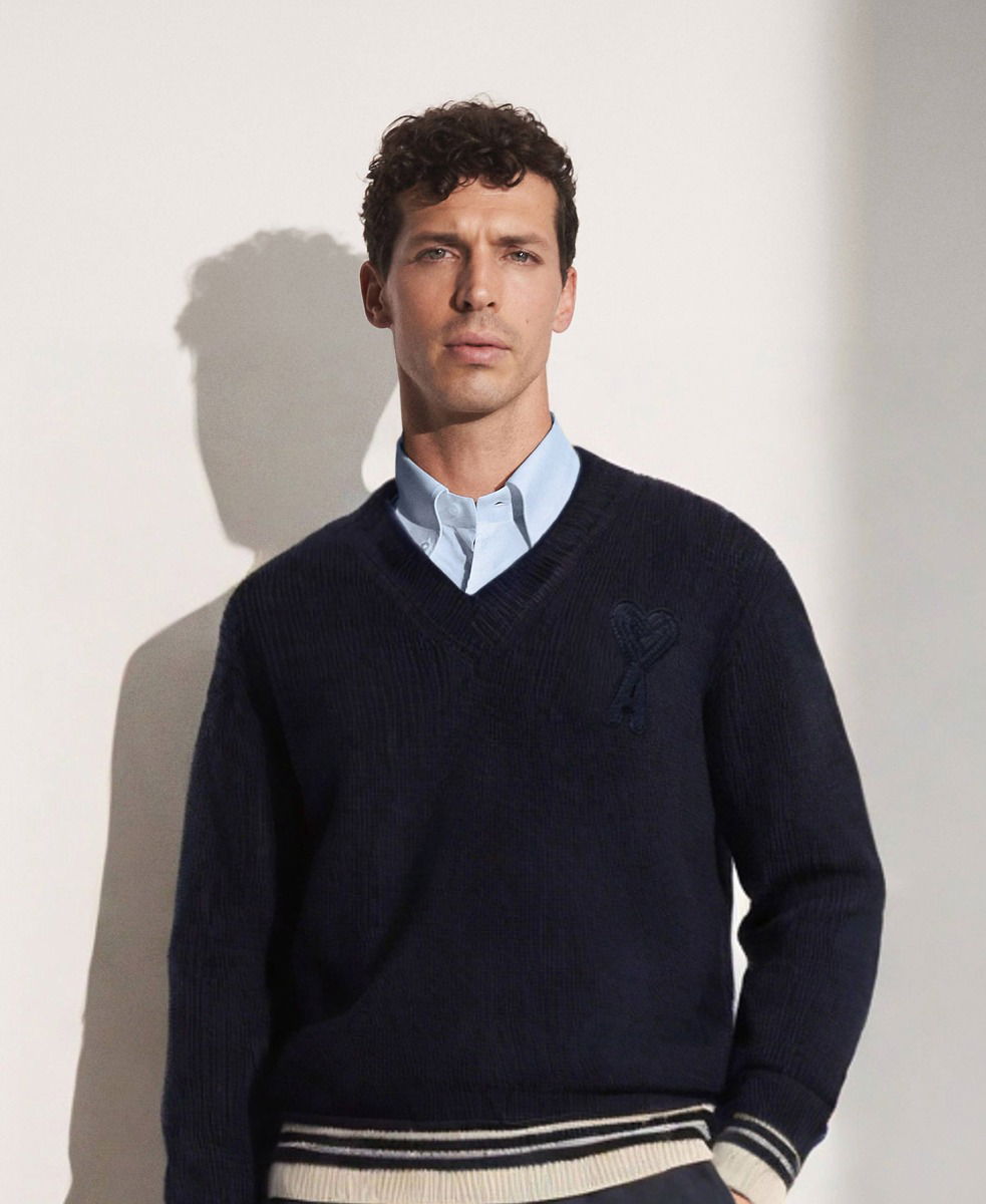 Mann mit lockigem Haar, hellblauem Hemd unter dunkelblauem V-Neck-Pullover mit Herz-Logo und gestreiftem Saum.