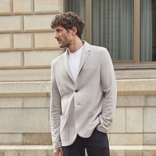 Mann mit hellgrau-beigem Sakko, weißem T-Shirt und schwarzer Hose steht vor hellbeigem Steinmauer-Hintergrund und schaut seitlich.