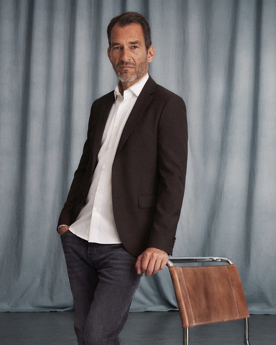 Mann mit offenem dunkelbraunem Sakko, weißem Hemd und grauer Jeans lehnt an braunem Stuhl vor blauem Vorhang. Mann mit offenem dunkelbraunem Sakko, weißem Hemd und grauer Jeans lehnt an braunem Stuhl vor blauem Vorhang.