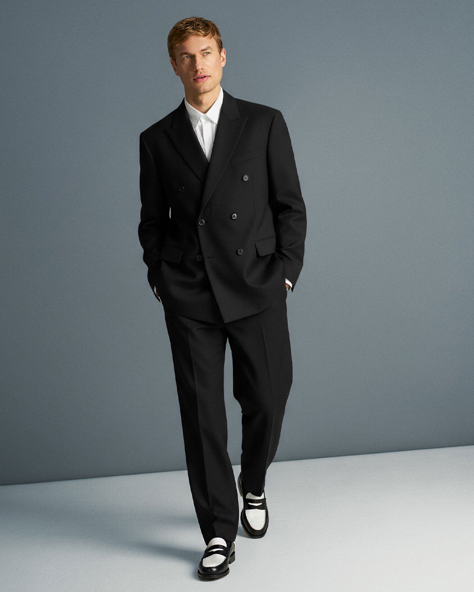 Mann trägt schwarzen oversized Anzug, weißes Hemd und schwarz-weiße Loafer, steht vor grauem Hintergrund.