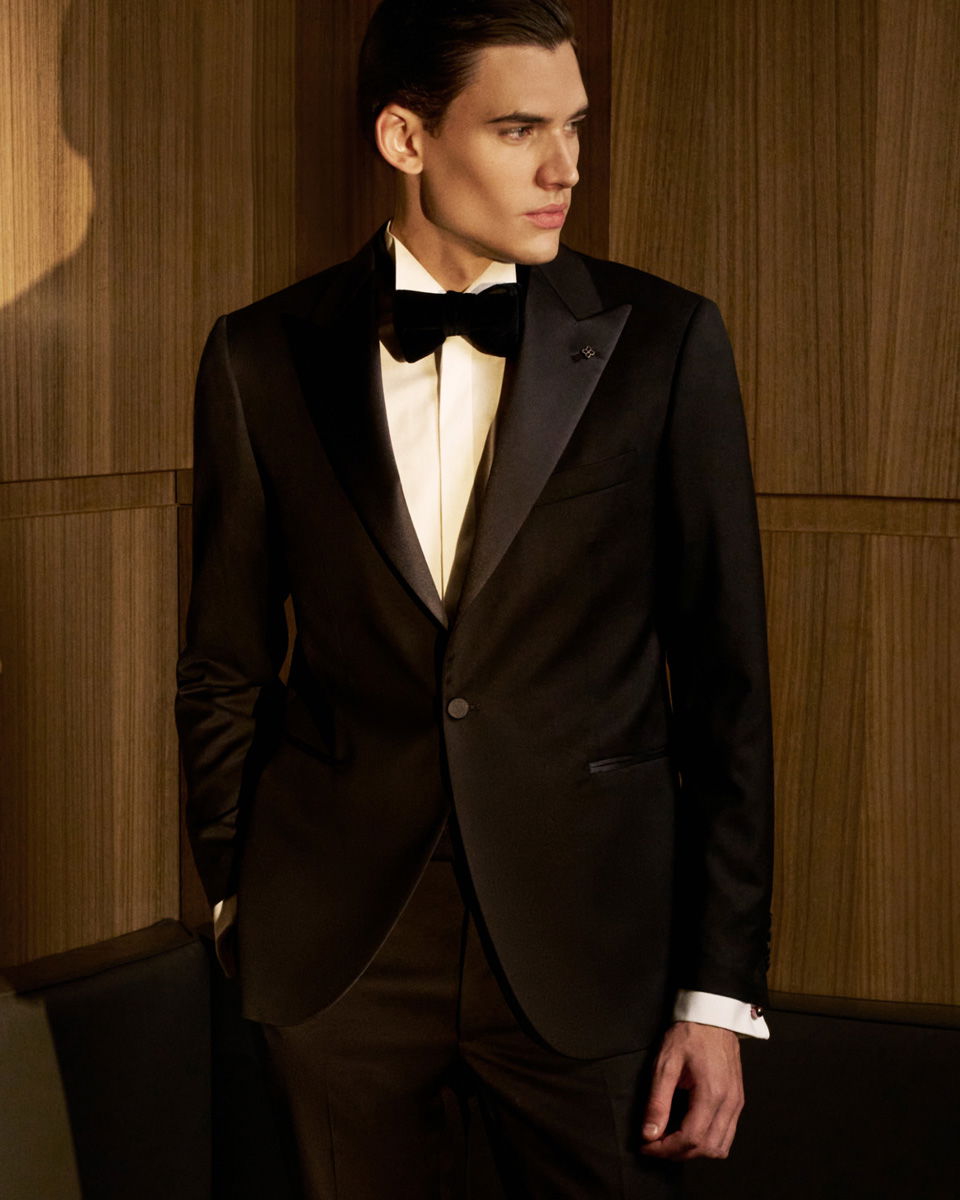 Junger Mann in schwarzem Smoking mit weißem Hemd und schwarzer Fliege steht vor Holzpaneelen, Blick nach rechts. Junger Mann in schwarzem Smoking mit weißem Hemd und schwarzer Fliege steht vor Holzpaneelen, Blick nach rechts.