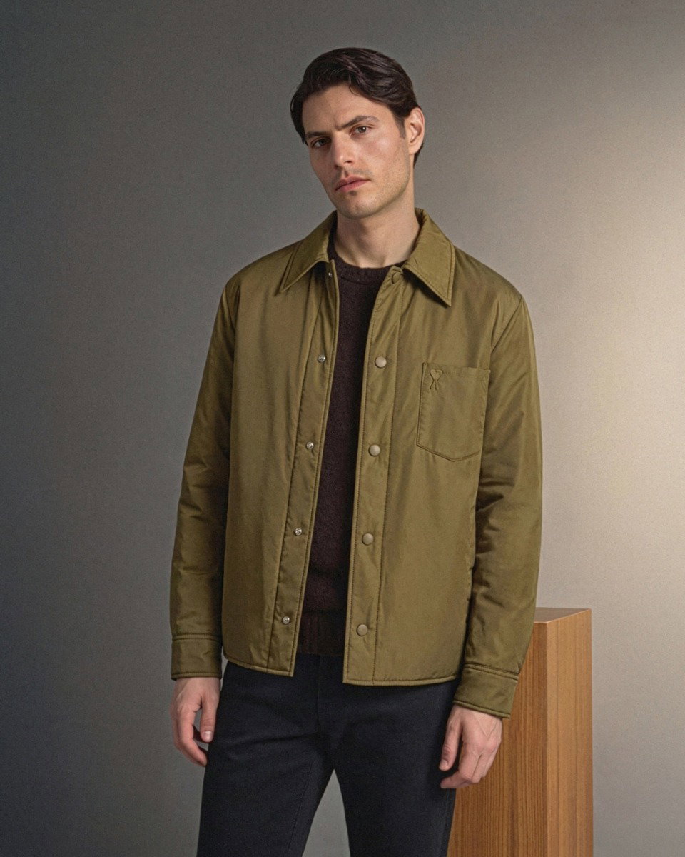 Mann mit olivgrünem Overshirt, schwarzem Pullover und schwarzer Hose steht vor neutralem Hintergrund neben Holzpodest.