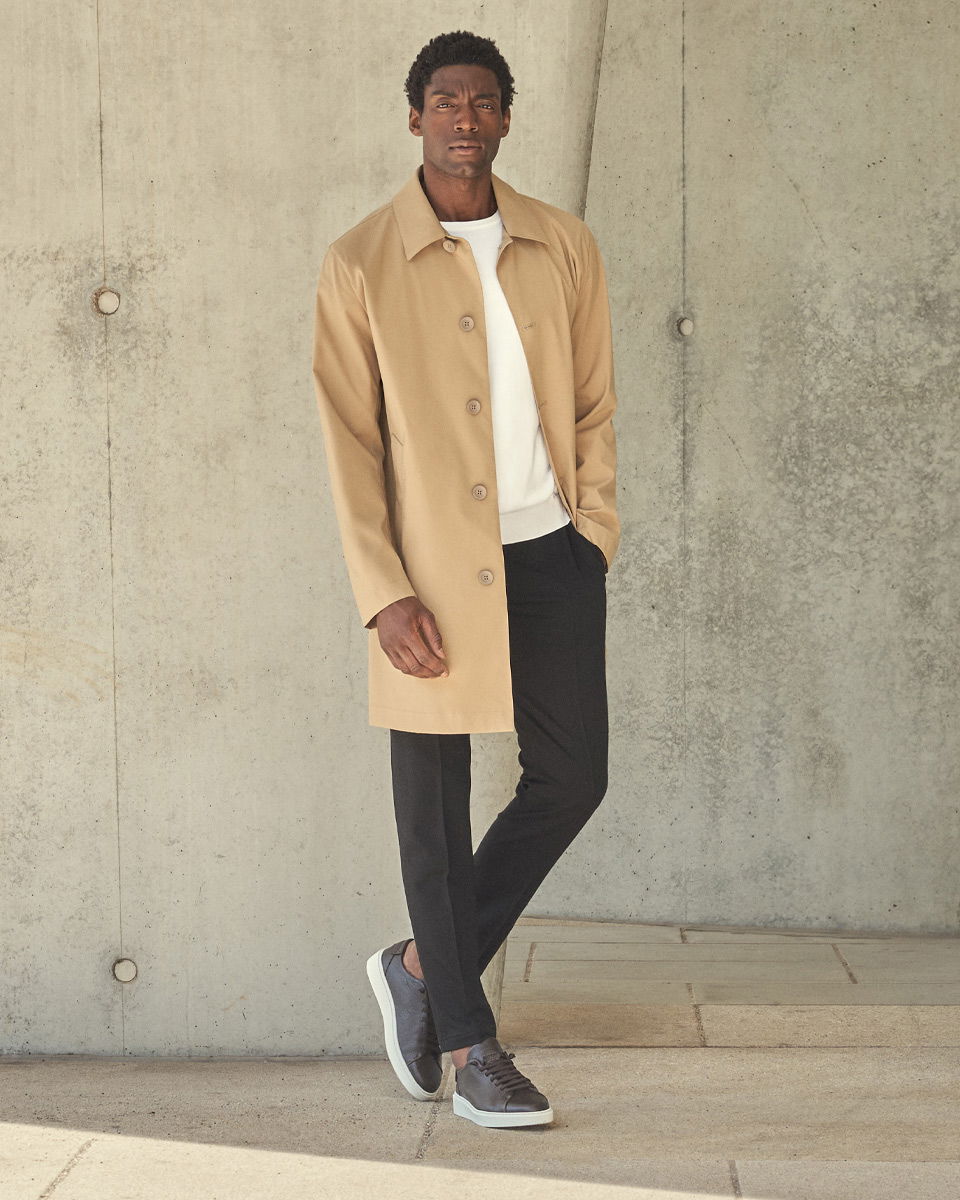 Mann trägt beigen Mantel, weißes Sweatshirt, schwarze Hose und dunkle Sneaker, steht vor grauer Betonwand. Mann trägt beigen Mantel, weißes Sweatshirt, schwarze Hose und dunkle Sneaker, steht vor grauer Betonwand.