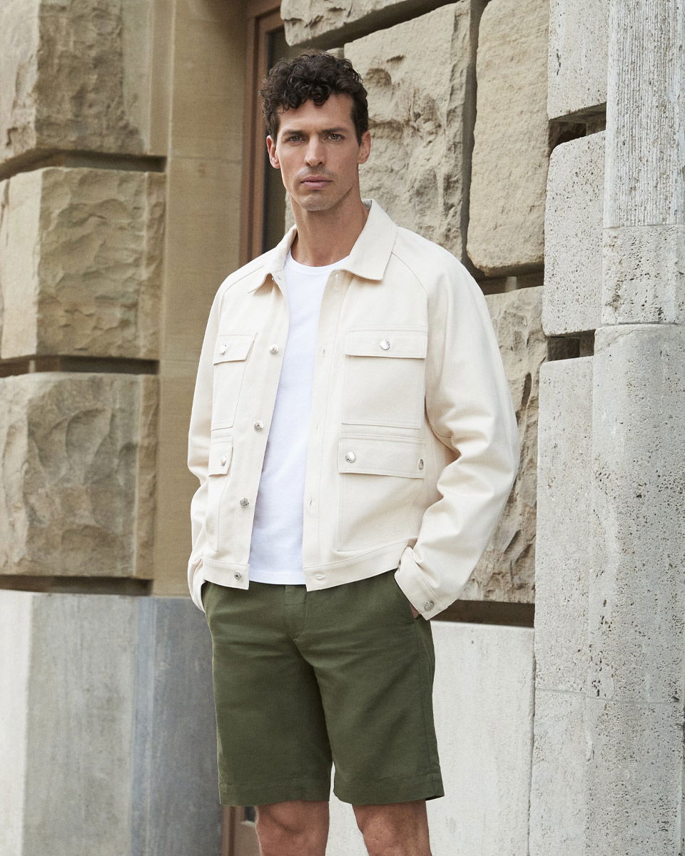 Mann mit weißer Jeansjacke, weißem T-Shirt und dunkelgrünen Shorts steht vor einer Steinwand mit Händen in den Taschen.