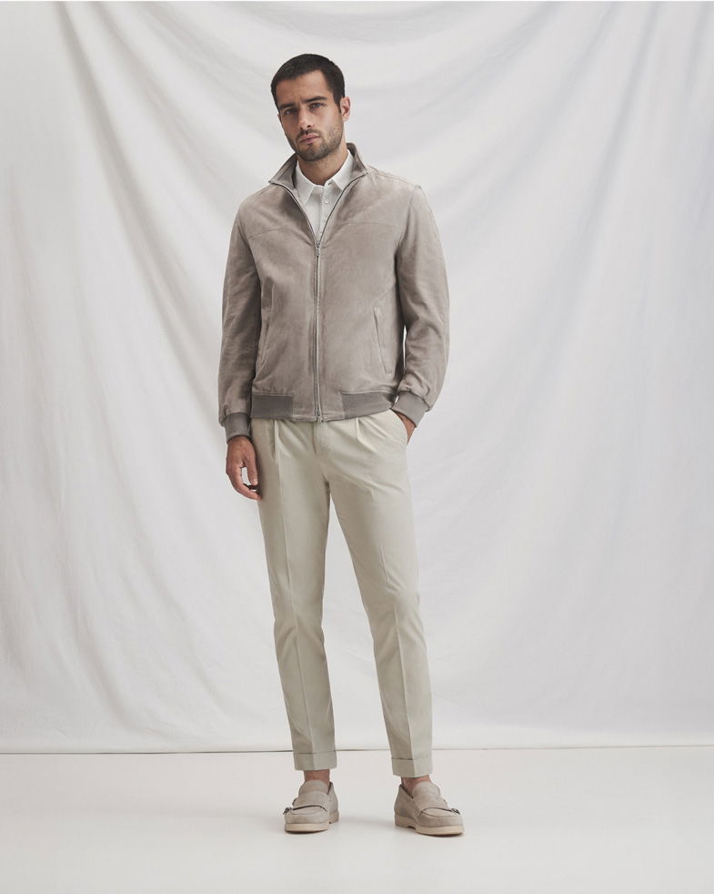 Mann trägt hellgraue Lederjacke über weißem Hemd, beige Chinohose und beige Loafers vor hellem Hintergrund.