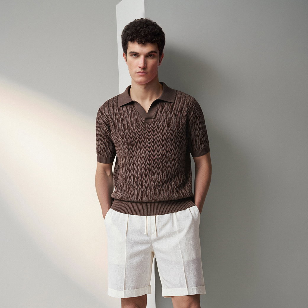 Mann trägt braunes kurzärmeliges Strickpolo mit Struktur und weiße Shorts, steht vor grau-weißem Hintergrund.