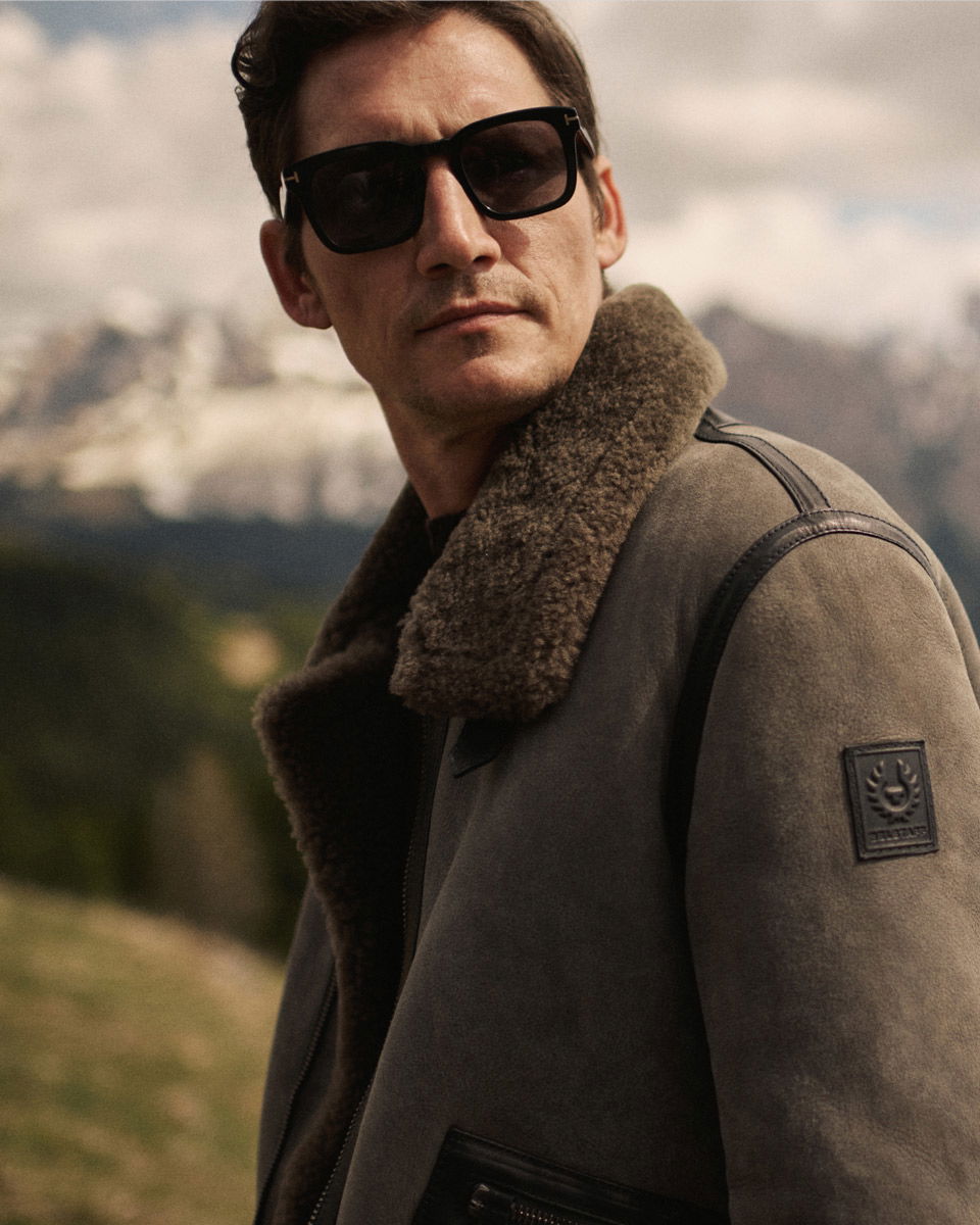Mann mit brauner Lederjacke mit Fellkragen und dunkler Sonnenbrille schaut nach hinten, Berge unscharf im Hintergrund. Mann mit brauner Lederjacke mit Fellkragen und dunkler Sonnenbrille schaut nach hinten, Berge unscharf im Hintergrund.