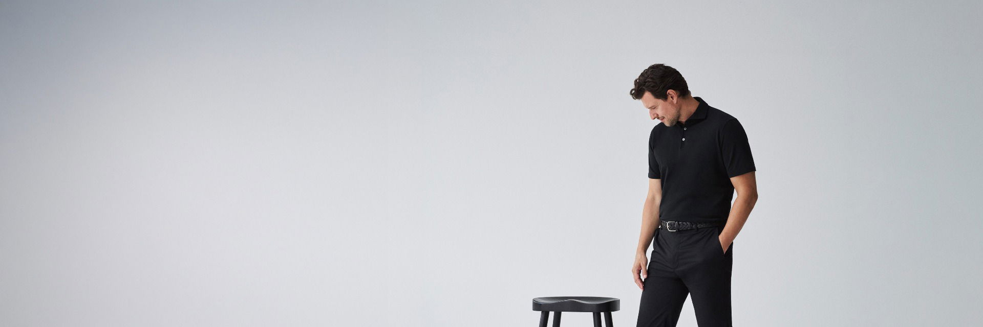 Mann in schwarzem Poloshirt und Hose mit schwarzem Ledergürtel steht vor grauem Hintergrund und schaut nach unten. Mann in schwarzem Poloshirt und Hose mit schwarzem Ledergürtel steht vor grauem Hintergrund und schaut nach unten.