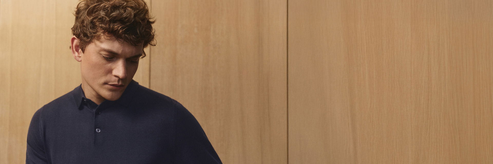 Junger Mann mit lockigen Haaren schaut nach unten, trägt ein dunkelblaues Langarm-Poloshirt aus Merinowolle vor Holzwand. Junger Mann mit lockigen Haaren schaut nach unten, trägt ein dunkelblaues Langarm-Poloshirt aus Merinowolle vor Holzwand.