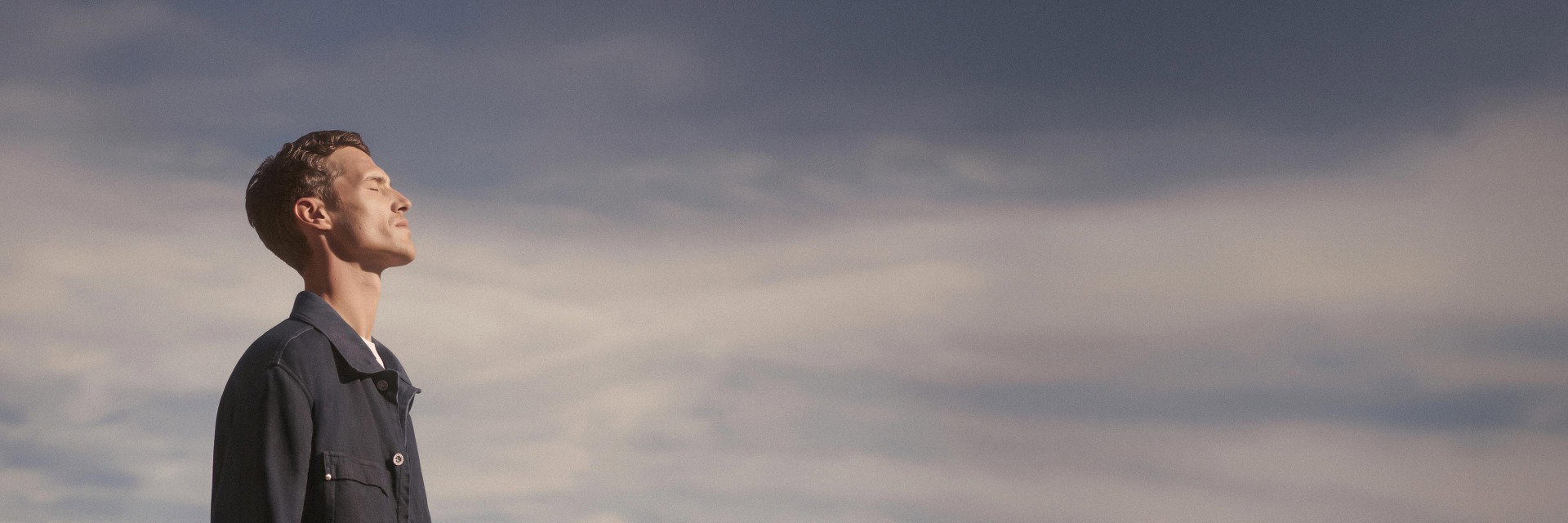 Mann mit geschlossenen Augen blickt entspannt zum Himmel, trägt dunkelblaues Overshirt vor wolkigem Himmel. Mann mit geschlossenen Augen blickt entspannt zum Himmel, trägt dunkelblaues Overshirt vor wolkigem Himmel.